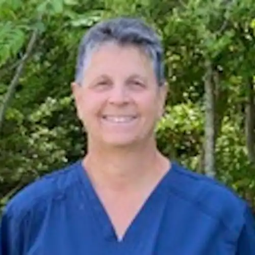 Elena, a physical therapist in scrubs, smiling outdoors with greenery in the background, representing her experience in home care therapy.