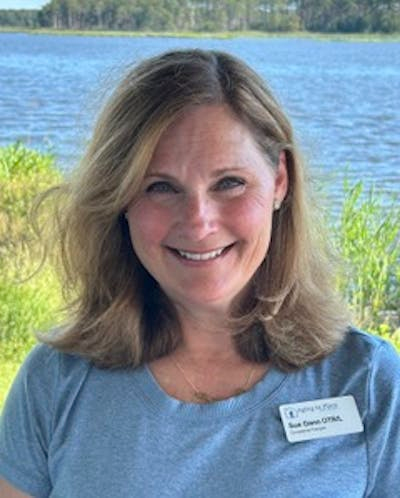 Sue Gann, OTR/L, smiling outdoors by a lake, wearing a name tag, showcasing her passion for occupational therapy and connection with seniors.