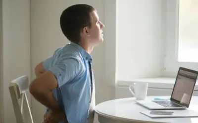 Man experiencing back pain while sitting at a table with a laptop and coffee cup, illustrating common issues related to physical health and wellness in aging.